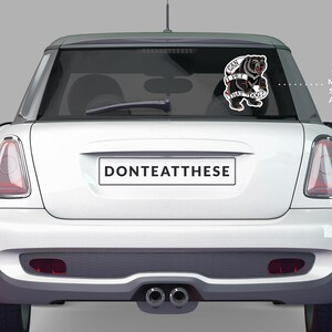 May include: A white Mini Cooper with a sticker on the rear window. The sticker features a black bear with the text "CAN I PET THAT DOG?" in a banner. The license plate reads "DONTEATTHESE".