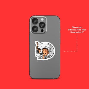 K&ouml;nnte beinhalten: Eine wei&szlig;e und graue Handyh&uuml;lle mit einem Aufkleber eines Comic-Katzen, die aus einem Sch&auml;del schaut. Gezeigt auf: iPhone 13 Pro Max. Gezeigte Gr&ouml;&szlig;e: 5 cm.