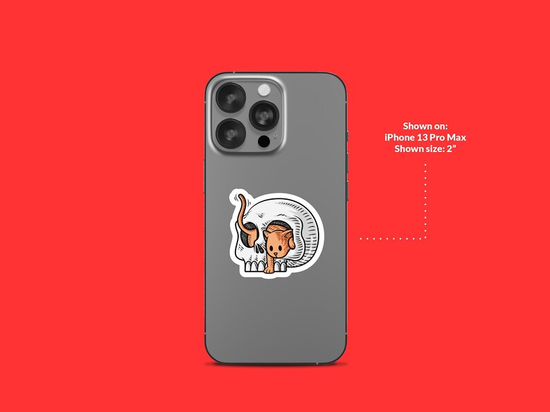 K&ouml;nnte beinhalten: Eine wei&szlig;e und graue Handyh&uuml;lle mit einem Aufkleber eines Comic-Katzen, die aus einem Sch&auml;del schaut. Gezeigt auf: iPhone 13 Pro Max. Gezeigte Gr&ouml;&szlig;e: 5 cm.