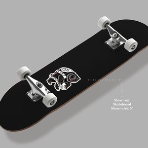May include: Black skateboard with white wheels and silver hardware. A sticker with a black dog and the text "CAN I PET THAT DOG?" is affixed to the board. The skateboard is shown on a gray background.