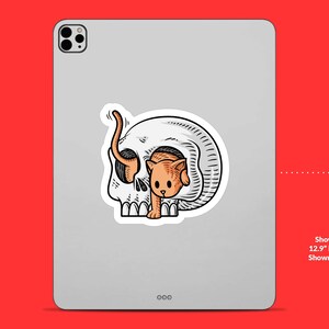 K&ouml;nnte beinhalten: Ein Aufkleber mit einer Cartoon-Illustration einer Katze, die aus einem Sch&auml;del schaut. Die Katze ist orange und wei&szlig;, der Sch&auml;del ist schwarz und wei&szlig;.