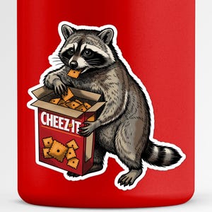 Peut inclure: Une surface rouge avec un autocollant représentant un raton laveur tenant une boîte rouge ouverte de crackers Cheez-It. Le raton laveur est gris et noir avec une queue rayée, et il mange un cracker orange carré tout en tenant la boîte.