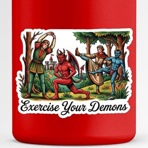 May include: Red mug with a sticker featuring a cartoon illustration of figures exercising in a forest. The sticker reads "Exercise Your Demons". The figures include a red devil, a blue demon, and two people in medieval clothing.