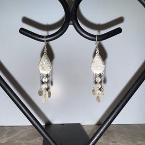 May include: A pair of silver-toned dangle earrings. Each earring features a textured, teardrop-shaped top with dangling chains and small, round charms. The earrings are displayed on a black metal stand.