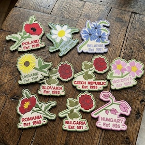 May include: Embroidered patches with floral designs and country names. Each patch shows a flower, the country's name, and the establishment year. Countries include Poland, Russia, Belarus, Ukraine, Slovakia, Czech Republic, Moldova, Romania, Bulgaria, and Hungary.