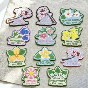 May include: A collection of twelve embroidered patches featuring various national flowers and country names with establishment years, including China, Japan, North Korea, Mongolia, South Korea, Thailand, Cambodia, Laos, Taiwan, Vietnam, and Philippines.