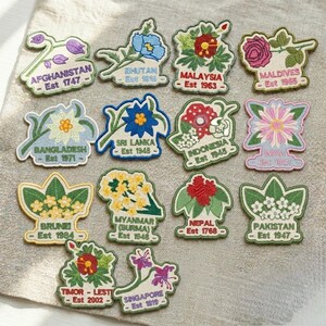 May include: A collection of fourteen embroidered patches featuring various national flowers and country names with their establishment years, including Afghanistan, Bhutan, Malaysia, Maldives, Bangladesh, Sri Lanka, Indonesia, India, Brunei, Myanmar, Nepal, Pakistan, Timor-Leste, and Singapore.