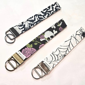 May include: Three fabric wrist keychains with Halloween patterns. One has bats, another a skull and roses, and the third a spiderweb. Each keychain has a metal ring and the text "Handmade by Victoria Mason".