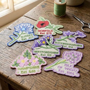 May include: A collection of embroidered floral patches, each representing a county such as Sussex, Norfolk, and Buckinghamshire. The patches display the county name and establishment year. They are arranged on a wooden table.