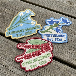 May include: Three embroidered patches with floral designs and text. The top left patch is light blue with white flowers and reads "DUNBARTONSHIRE Est 1890". The top right patch is blue with blue flowers and reads "PERTHSHIRE Est 1124". The bottom patch is red and green and reads "STIRLINGSHIRE Est 1124".