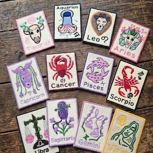 May include: A collection of embroidered zodiac sign patches, each with a unique design and colour scheme. Signs include Taurus, Aquarius, Leo, Aries, Capricorn, Cancer, Pisces, Scorpio, Libra, Gemini, Sagittarius, and Virgo, with their names displayed.