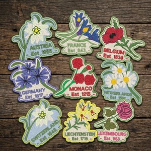 May include: Nine embroidered floral patches, each representing a different country. The patches feature various flowers in vibrant colors, with the country name and establishment year embroidered below each flower. The background is a rustic wooden surface.