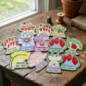 May include: A collection of embroidered fabric patches featuring various South American national flowers and country names with establishment dates, including Brazil, Argentina, Chile, Colombia, Peru, Ecuador, Bolivia, Paraguay, Venezuela, Guyana, Suriname, and Uruguay.