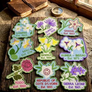 May include: Nine embroidered patches featuring various national flowers and country names including Gambia Est 1965, Guinea Est 1958, Ghana Est 1957, Mali Est 1235, Nigeria Est 1960, Guinea-Bissau Est 1973, Togo Est 1960, Cote D'Ivoire Est 1893, and Sierra Leone Est 1961.