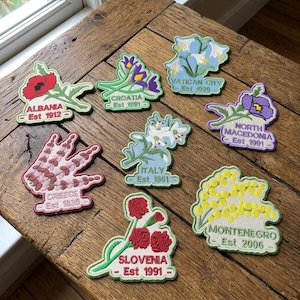 May include: Embroidered patches featuring floral designs and country names, including Albania, Croatia, Vatican City, Greece, Italy, North Macedonia, Slovenia, and Montenegro. Each patch has the country name and establishment year.