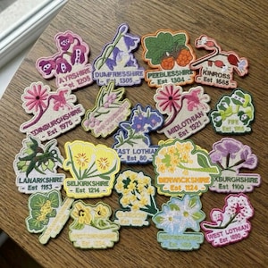 May include: A collection of embroidered fabric patches featuring various floral designs and Scottish county names with establishment dates, including Ayrshire, Dumfriesshire, Peeblesshire, Kinross, Edinburghshire, Midlothian, Fife, Lanarkshire, Selkirkshire, and others.