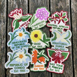 May include: A collection of nine embroidered patches featuring various national flowers and country names including Angola, Cameroon, Central African Republic, Democratic Republic of the Congo, Chad, Equatorial Guinea, Republic of Congo, Gabon, and Sao Tome & Principe.