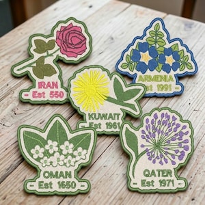 May include: Five embroidered fabric patches featuring floral designs and country names. The patches depict a pink rose for Iran Est 550, blue flowers for Armenia Est 1991, a yellow dandelion for Kuwait Est 1961, white flowers for Oman Est 1650, and purple allium for Qatar Est 1971.
