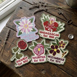 May include: Five embroidered fabric patches featuring floral designs and country names: Malawi Est 1964, Madagascar Est 1960, Rwanda Est 1962, Mauritius Est 1638, and Mozambique Est 1975. Each patch has a distinct flower and green leaves on a light background.