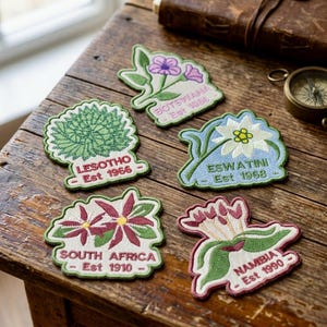 May include: Five embroidered fabric patches featuring botanical illustrations and country names. The patches include Botswana, Lesotho, Eswatini, South Africa, and Namibia, each with their respective establishment dates stitched in colorful thread.