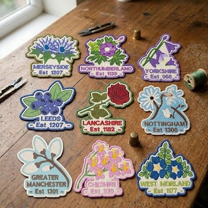 May include: A selection of embroidered patches, each depicting a county flower and name. The patches include designs for Merseyside, Yorkshire, and Lancashire, among others. The patches are displayed on a wooden table.