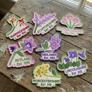 May include: A collection of embroidered floral patches, each representing a county such as Warwickshire, Staffordshire, and Lincolnshire. Each patch includes a floral design, county name, and establishment year.