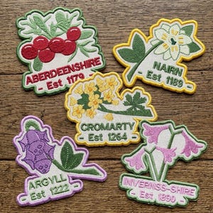May include: A collection of embroidered patches with floral and botanical designs, each featuring a place name and establishment date. The patches showcase images of berries, flowers, and leaves in colors like red, yellow, and purple. Text includes "ABERDEENSHIRE", "NAIRN", "CROMARTY", "ARGYLL", and "INVERNESS-SHIRE".