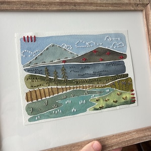 May include: A framed watercolor painting of a landscape with mountains, a lake, and trees. The painting is stitched with colorful thread, creating a textured effect.
