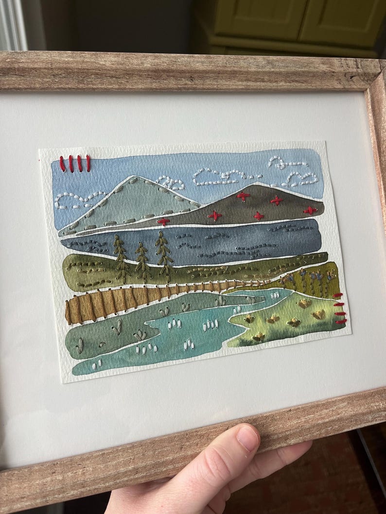 May include: A framed watercolor painting of a landscape with mountains, a lake, and trees. The painting is stitched with colorful thread, creating a textured effect.