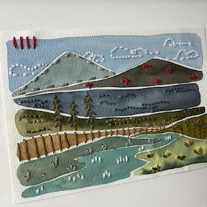 May include: A watercolor and embroidery artwork depicting a landscape with mountains, a lake, and trees. The sky is blue with white clouds, and the mountains are green and brown. The lake is blue and green, and the trees are green. The artwork is framed in a wooden frame.