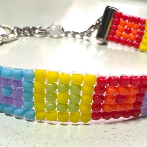 May include: A colorful beaded bracelet with a rainbow pattern. The bracelet features a variety of colors, including red, orange, yellow, green, blue, and purple. It has a silver clasp and is approximately 0.75 inches wide.
