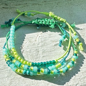 May include: A multi-strand bracelet with green and blue beads and green string. The bracelet is made of multiple strands of string with beads woven in.