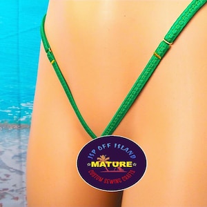 Puede incluir: Primer plano de un tanga bikini verde. Las correas son finas y ajustables, con herrajes dorados. El bikini está hecho de una tela brillante y lisa. Un logotipo circular con el texto "JSP OFF ISLAND MATURE CUSTOM SEWING CRAFTS" es visible.