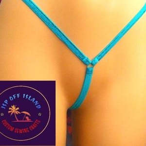 May include: A turquoise thong bikini bottom. The bikini bottom has thin straps and a ring detail at the front. The image also includes a logo with the text "JSP OFF ISLAND CUSTOM SEWING CRAFTS".