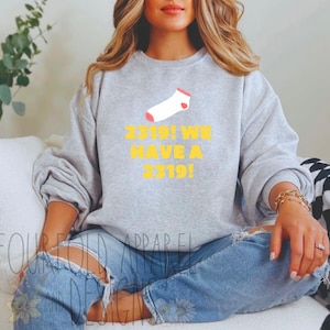 Op de afbeelding: Een grijs sweatshirt met een gele afbeelding waarop staat "2319! We hebben een 2319!" en een roze sok afbeelding.