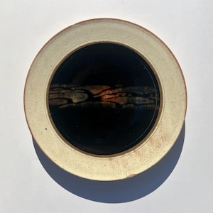 May include: A white ceramic plate with a brown rim and a black centre. The centre has a brown and orange design.