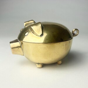 May include: A brass piggy bank shaped like a pig. It has a hinged lid, small legs, and a curled tail. The ears and snout are clearly defined. The bank is made of shiny, polished brass.
