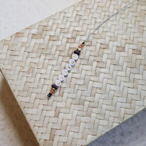 May include: A beaded keychain spelling out the word "CUSTOM" in gold letters on white beads. The keychain features brown and black accent beads and is displayed on a woven, light-colored surface.