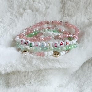 Może przedstawiać: Zestaw bransoletek z koralików w odcieniach różu, zieleni i bieli. Jedna bransoletka wymawia imię "SHELLYEAH" za pomocą kolorowych koralików z literami. Złote gwiazdki i zawieszki dodają szczegółów bransoletkom.