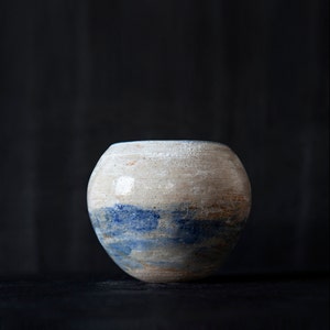 Op de afbeelding: Een ronde keramische vaas met een witte en blauwe glazuur. De vaas heeft een rustieke, handgemaakte uitstraling.