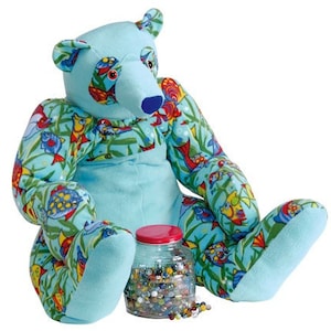 May include: A blue plush teddy bear with a colorful fish print pattern. The bear is sitting with its legs crossed and a glass jar filled with colorful marbles is in front of it.