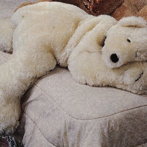 May include: A large, white, plush polar bear stuffed animal is lying on a bed with its head resting on its paws. The bear is made of soft, fluffy material and has a realistic appearance.