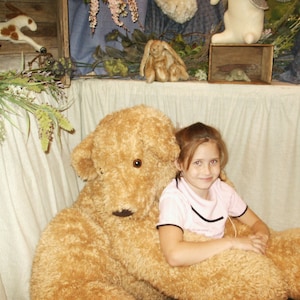 May include: A large, plush, golden-brown teddy bear with a smiling girl sitting on its lap. The bear has a textured fur appearance and a friendly expression. The background includes decorative elements like flowers and a wooden box.