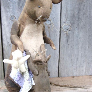 Pode incluir: Uma boneca de rato de tecido castanho feita à mão com barriga bege clara e orelhas grandes. O rato segura um pequeno urso branco e um tecido com estampa floral. Uma boneca de rato menor se agarra ao seu lado.