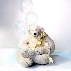 May include: A plush, cream-colored teddy bear with a black nose and stitched paws. It has a cream-colored ribbon tied around its neck and is holding three clear balloons. The bear is sitting on a white surface.
