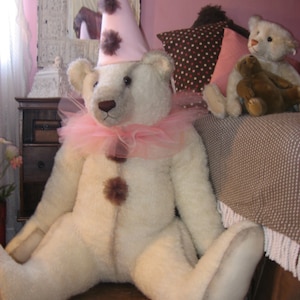 May include: A large white teddy bear wearing a pink and white tutu and a pink and white cone-shaped hat with black pom-poms. The bear is sitting on a bed with a brown and white patterned pillow and a pink blanket.