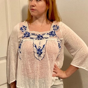 May include: White sheer blouse with blue floral embroidery. The embroidery features a delicate floral pattern with blue and white accents. The blouse has a square neckline and 3/4 sleeves.