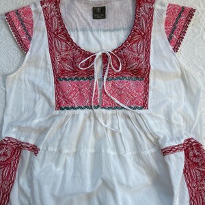 Op de afbeelding: Witte katoenen blouse met rode en groene borduursels. De blouse heeft een geplooide halslijn en korte mouwen.