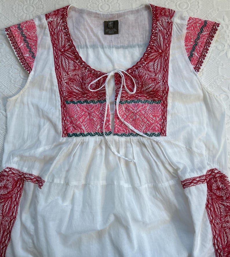 Op de afbeelding: Witte katoenen blouse met rode en groene borduursels. De blouse heeft een geplooide halslijn en korte mouwen.