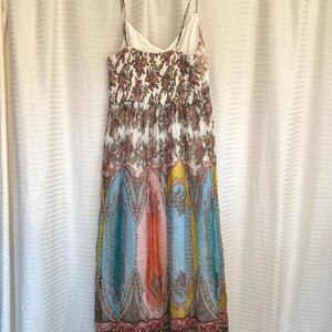 May include: A colorful, patterned maxi dress with spaghetti straps. The dress features a floral bodice and a skirt with vertical panels of blue, pink, yellow, and orange paisley designs. The dress is hanging on a white hanger against a white curtain.